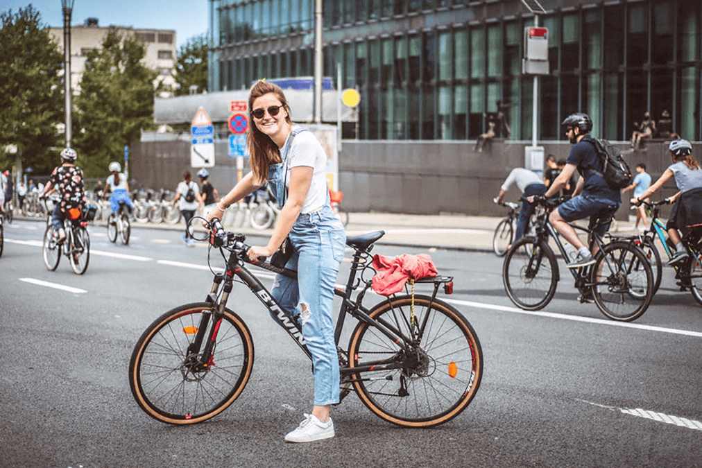 Vrouw met zonnebril zit op de fiets en kijkt naar de camera. Achter haar rijden er heel wat fietsers voorbij.
