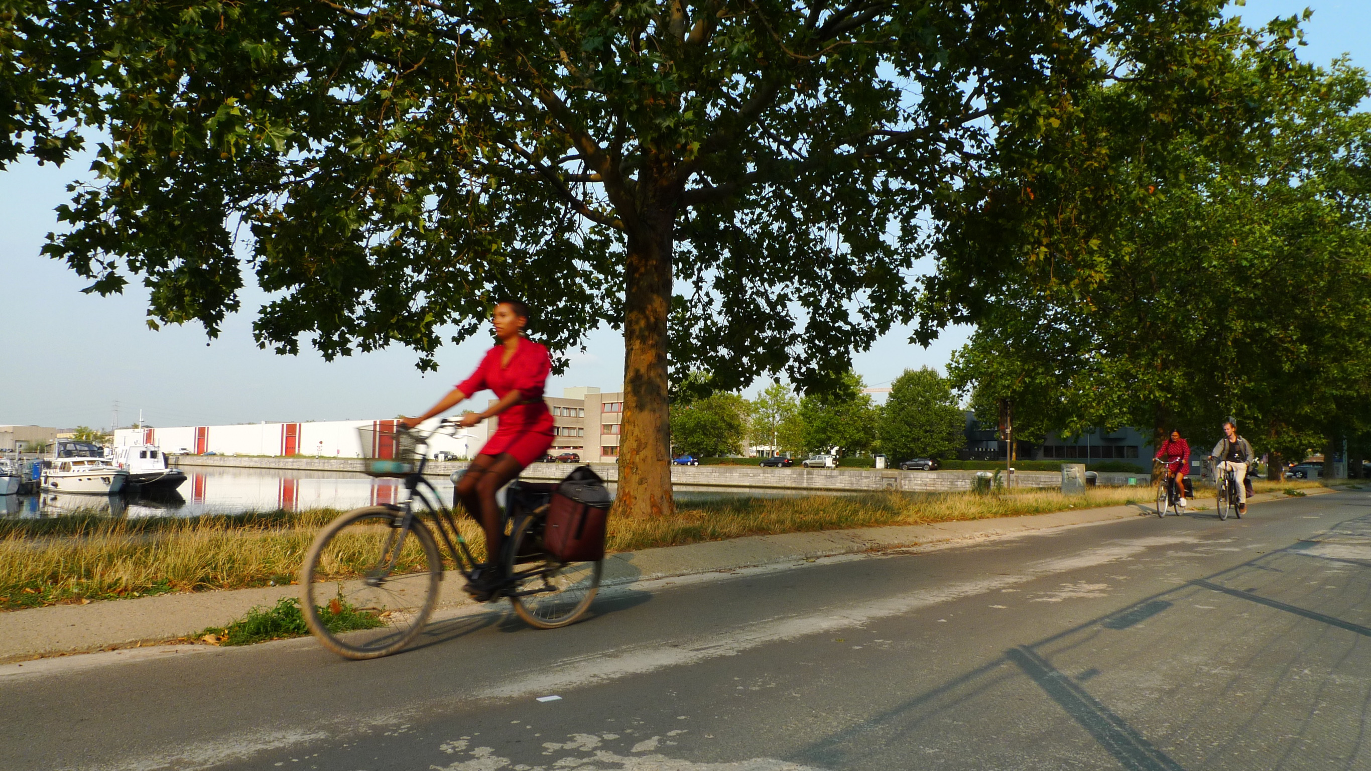 Een fietspad op een zonnige dag. Een vrouw in een rode jurk fietst snel voorbij, richting de camera. Verderop fietsen twee fietsers weg van de camera. 