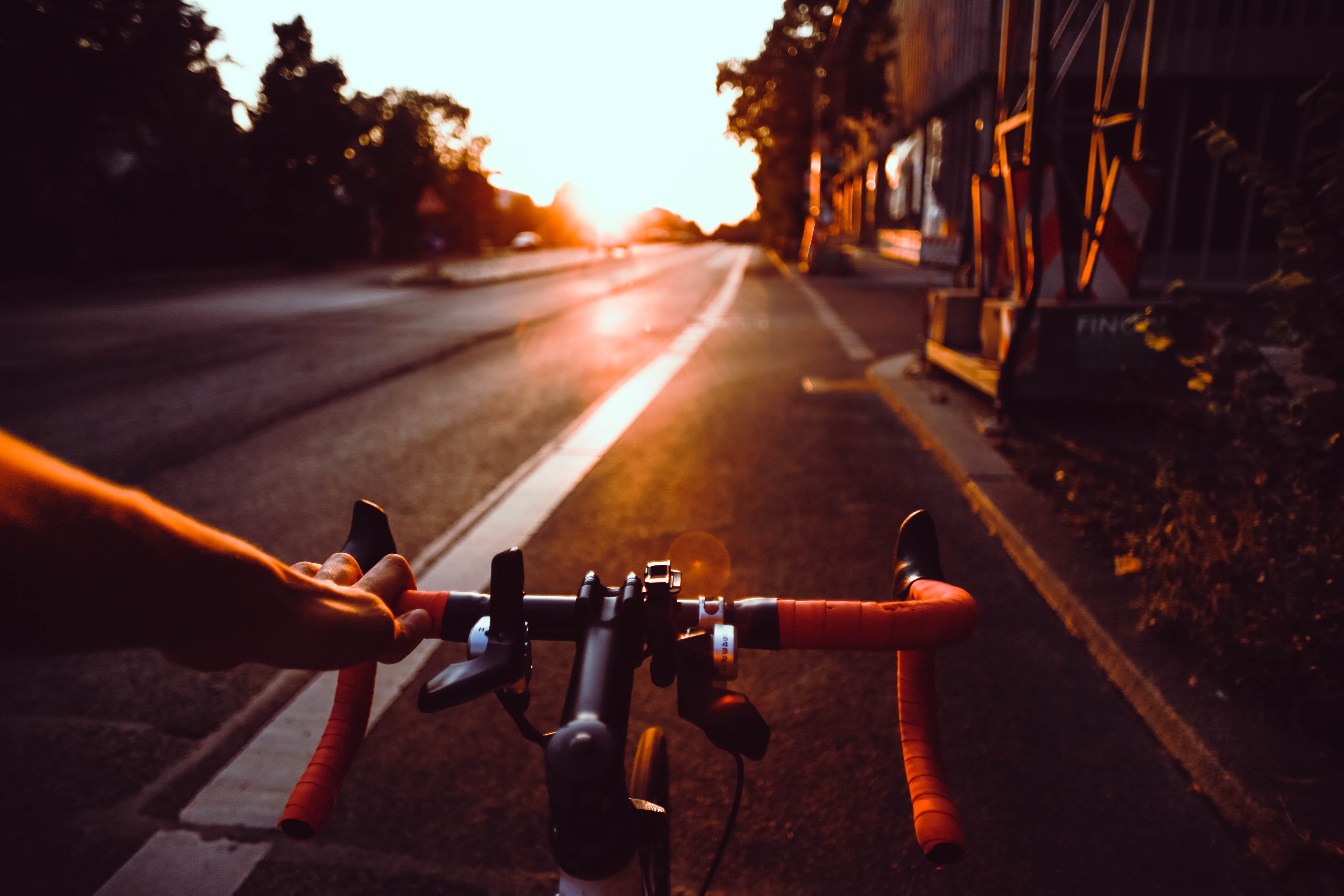 Het fietsstuur van een sportieve fiets met een hand aan het stuur. Ervoor het fietspad met bomen erlangs en de zon die opkomt. De sfeer is zomers.