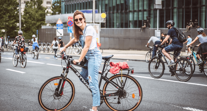 Vrouw met zonnebril zit op de fiets en kijkt naar de camera. Achter haar rijden er heel wat fietsers voorbij.