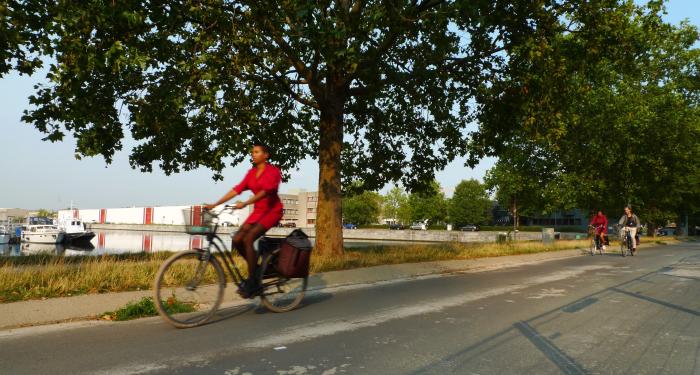 Een fietspad op een zonnige dag. Een vrouw in een rode jurk fietst snel voorbij. Verderop fietsen twee fietsers weg van de camera. 