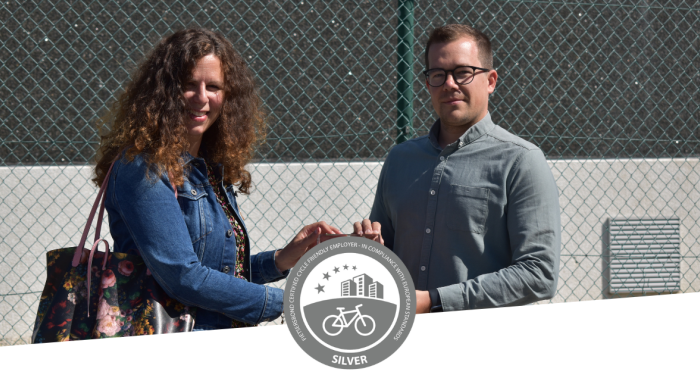 Een foto van Cindy van de Fietsersbond die een award overhandigt aan Geert Hennebel. De onderkant van de foto is bedekt met een witte band en het logo van een zilveren Cycle Friendly Employer Certificate.  